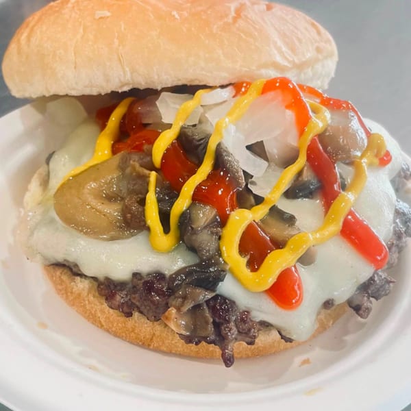 Cheesesteak sandwich with melted cheese, mushrooms, onions, ketchup, and mustard on a white plate.