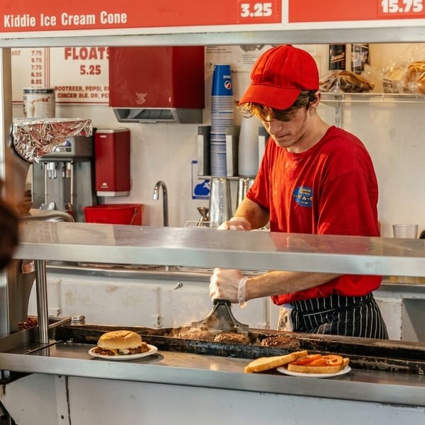 A cook in a red shirt and cap grills burgers behind a counter with menu boards above.