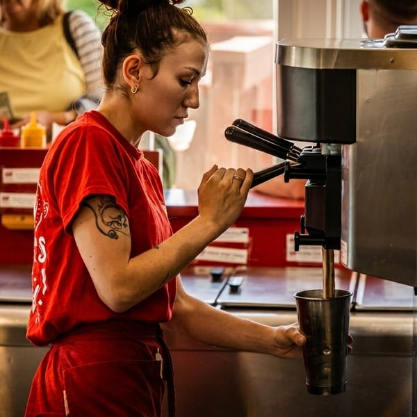 A woman in a red shirt and apron dispenses a drink from a machine into a metal cup.